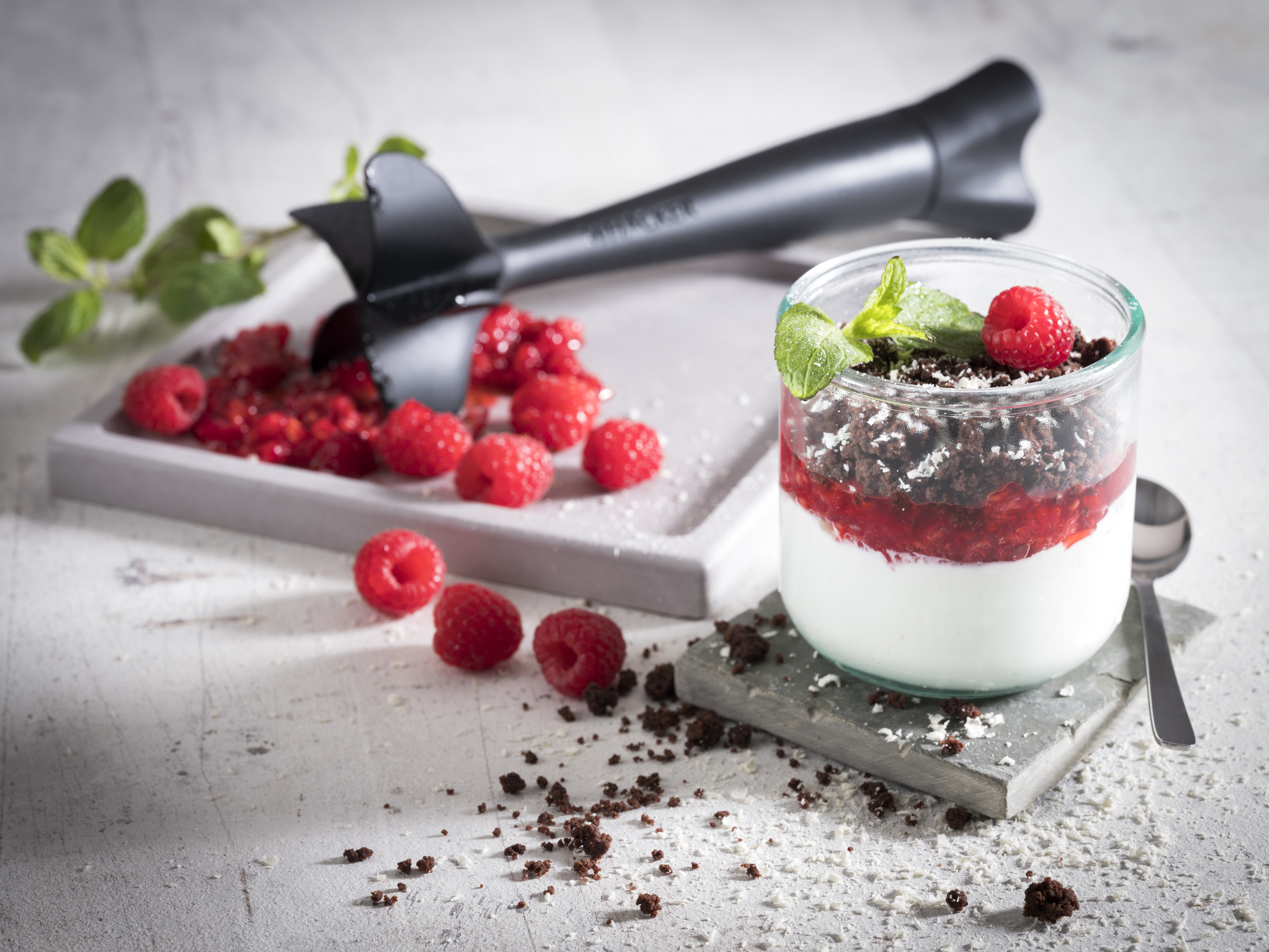 Mascarpone-Limetten-Creme mit Himbeeren und Schokoladencrumble Ein Dessert im Glas mit Schichten aus Joghurt, Himbeeren und Schokoladencrumble, dekoriert mit Minzblättern, auf einem Tisch neben frischen Himbeeren und einem Stampfer.