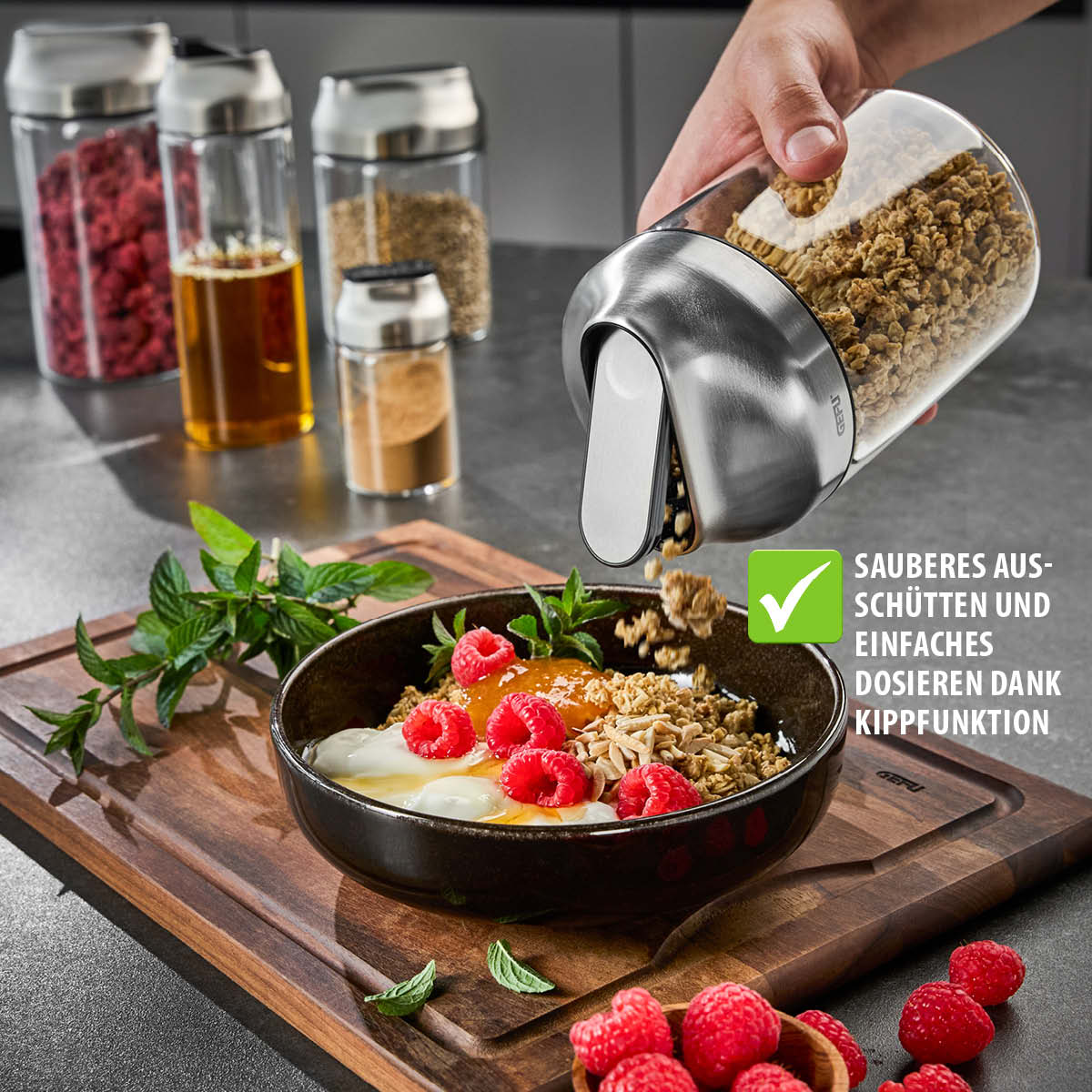 Hand kippt Granola aus einem Spender auf Schüssel mit Joghurt und Beeren, umgeben von Vorratsgläsern