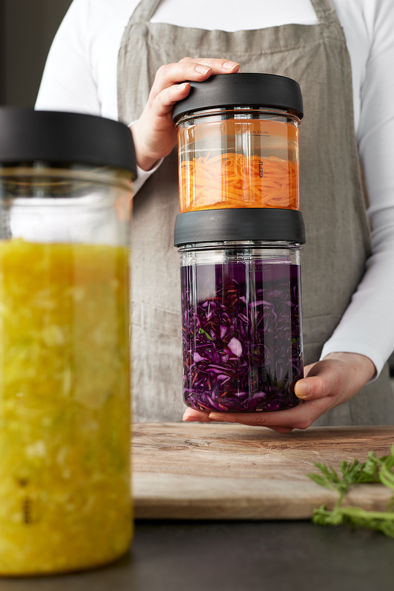 Fermentiergläser mit eingelegtem Gemüse in verschiedenen Farben Eine Person in einer Schürze hält zwei Fermentiergläser mit eingelegtem orangefarbenem und violettem Gemüse, im Vordergrund steht ein weiteres Glas mit gelbem Inhalt.