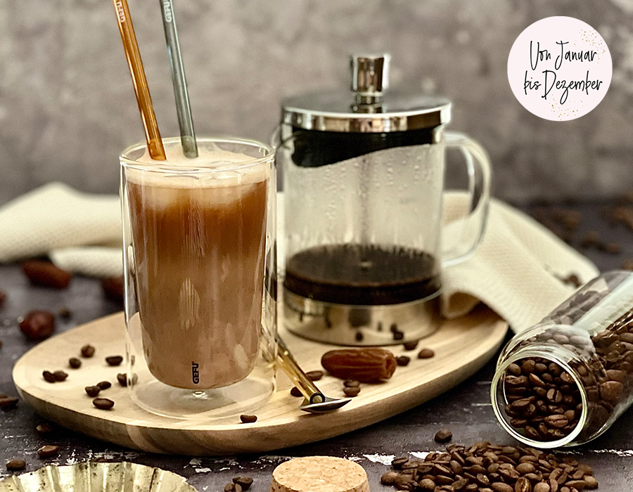 Protein Eiskaffee Ein Glasbecher mit Latte Macchiato auf einem Tablett, daneben eine French Press und ein Glas mit Kaffeebohnen.
