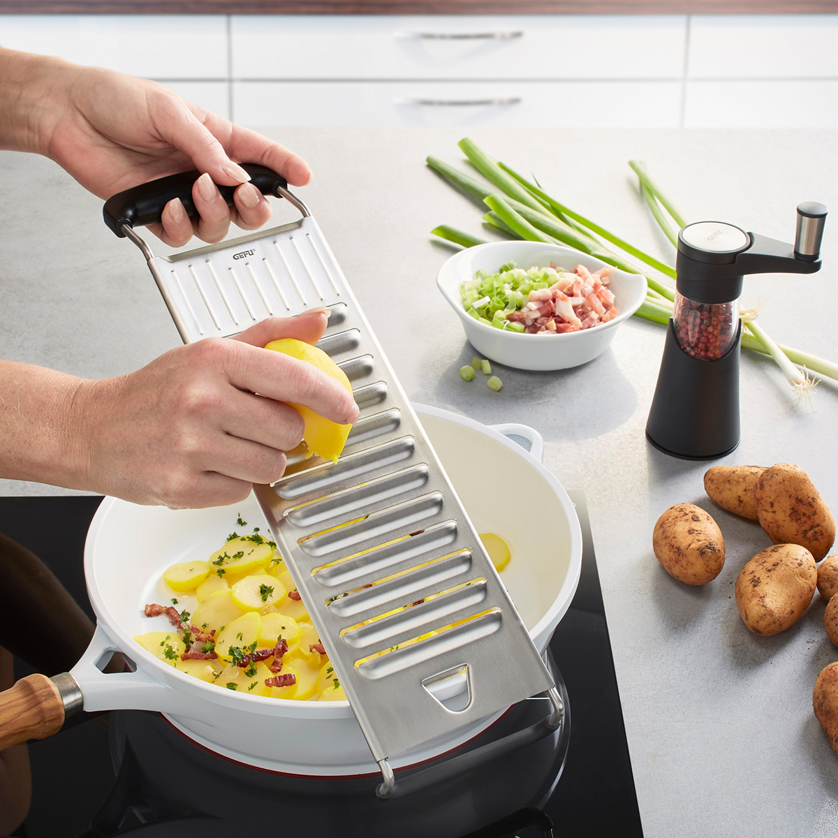 Kartoffelhobel in Benutzung auf einer modernen Küchenarbeitsplatte Eine Person reibt mit einem Kartoffelhobel Kartoffeln in eine weiße Pfanne. Im Hintergrund sind Frühlingszwiebeln, eine Schale mit Speck und Frühlingszwiebeln, ein Pfeffermühlenstreuer und ungekochte Kartoffeln sichtbar.