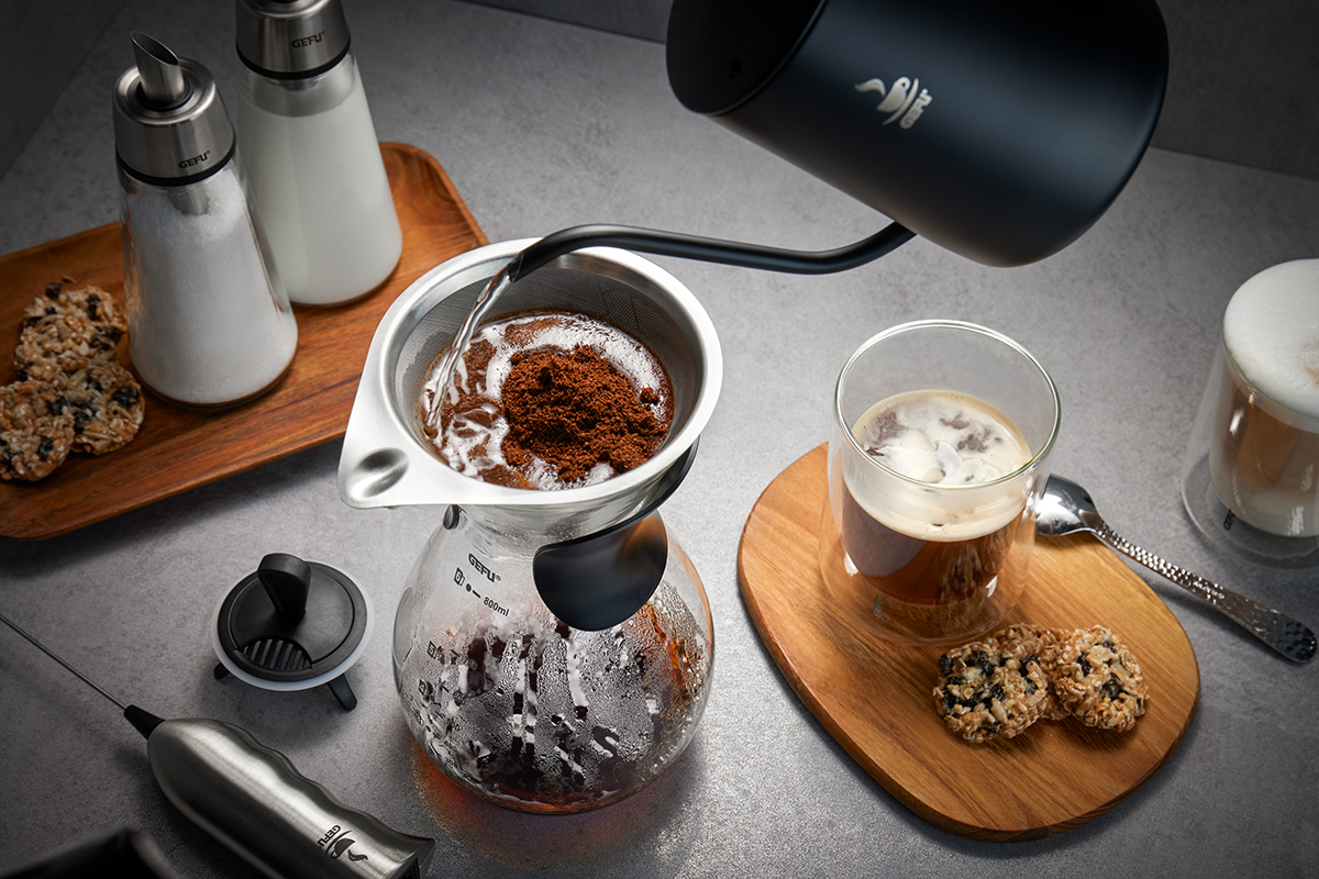 Eine stilvolle Kaffeezubereitung mit Handfilter auf einer Kanne, begleitet von einem Glas Kaffee und Keksen auf einem Tisch. Im Hintergrund stehen Milch und Zucker.