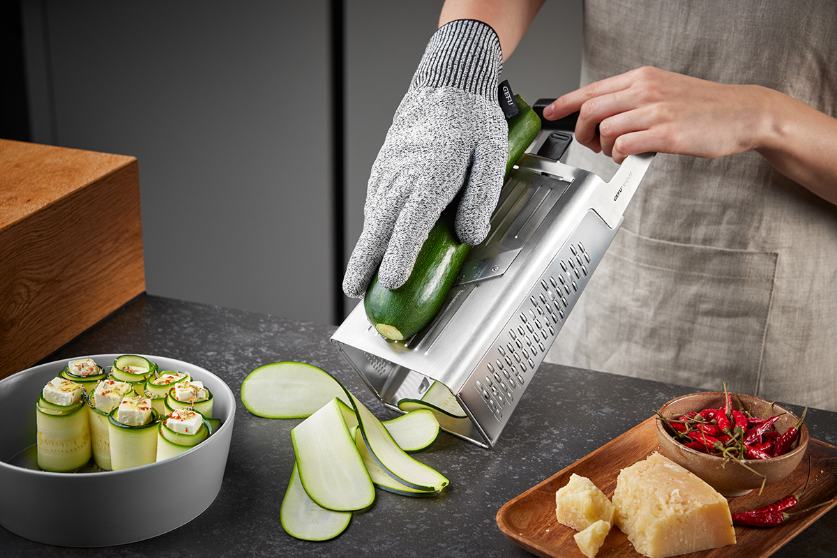 Küchenhobel mit Zucchini und Sicherheits-Handschuh Person verwendet einen Küchenhobel zum Schneiden einer Zucchini, während sie einen Sicherheits-Handschuh trägt. Im Vordergrund liegen geschnittene Zucchinistreifen, fertig angerichtete Zucchiniröllchen in einer Schale und Gewürze auf einem Brettchen.